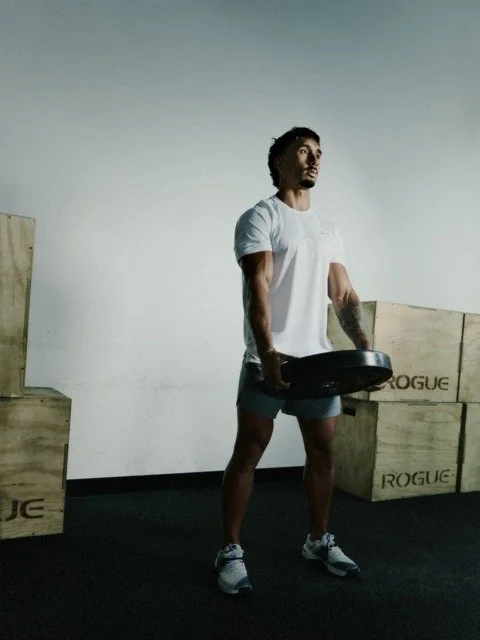 A man performing a standing weightlifting exercise with a weight plate in a gym, surrounded by wooden plyometric boxes.