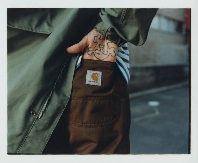 Close-up of a person wearing a green jacket and brown Carhartt pants with hand in pocket, revealing tattooed wrist and part of a striped shirt.