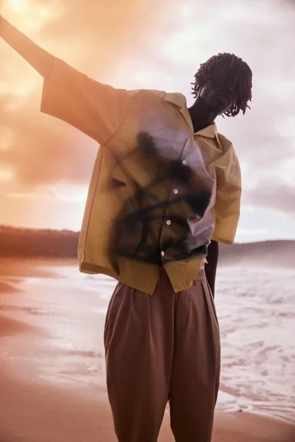 Person standing on the beach during sunset, wearing a yellow short-sleeve button-up shirt and brown pants, with one arm raised and looking down.