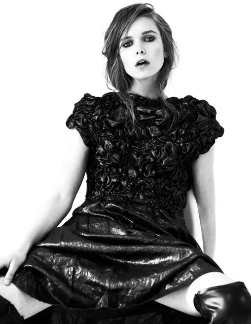 Black and white photo of a young woman with dark eye makeup and messy hair, wearing a textured, puff-sleeve top and a shiny, patent leather skirt, sitting with one leg up.