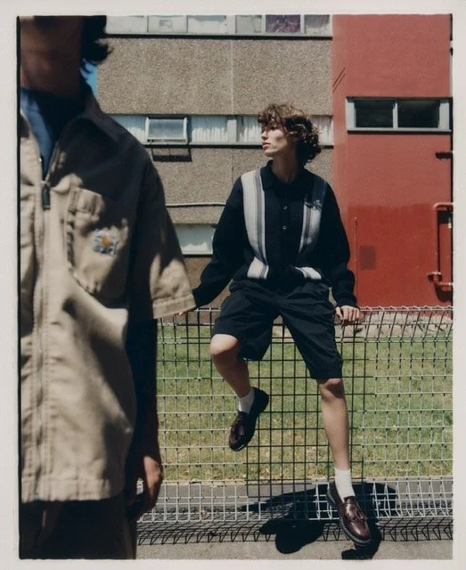 A young man with curly hair wearing a black and white varsity jacket, black shorts, and brown shoes sitting on a metal fence in an urban area.