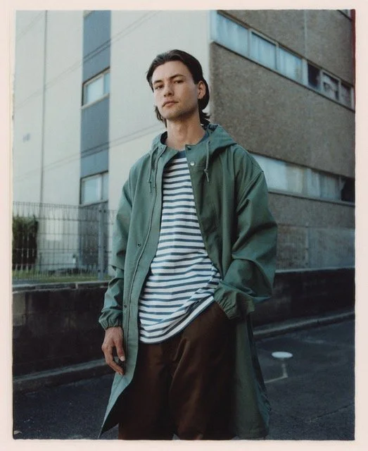 A young man with dark hair standing outdoors in front of an apartment building, wearing a green raincoat over a striped shirt and brown pants.