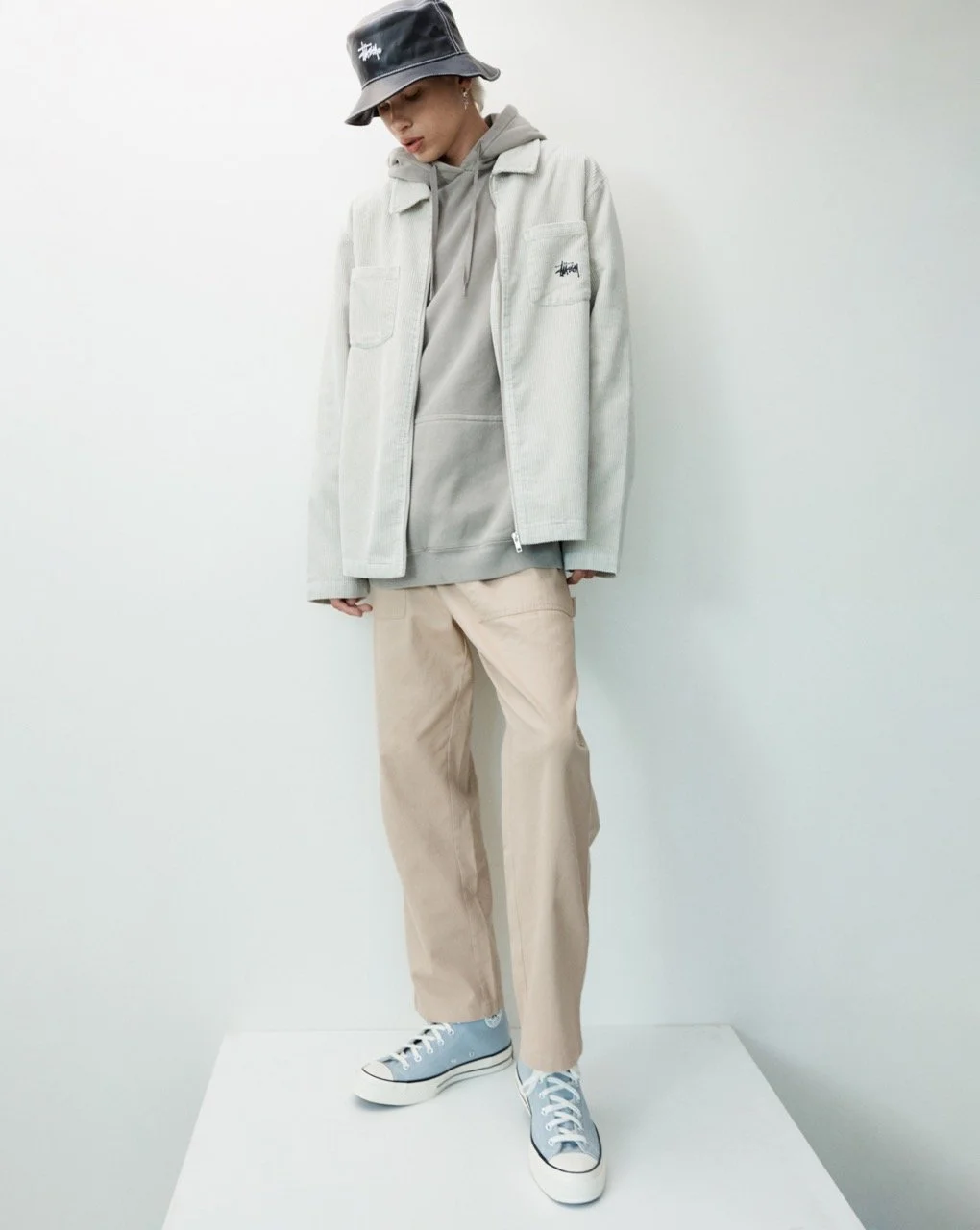 A person standing against a plain white background, dressed in a beige bucket hat, gray hoodie, light-colored jacket, beige pants, and blue sneakers.