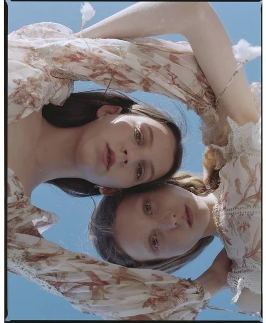 Two young women taking a selfie from below against a blue sky, with one resting her arm on her head and both looking down at the camera.