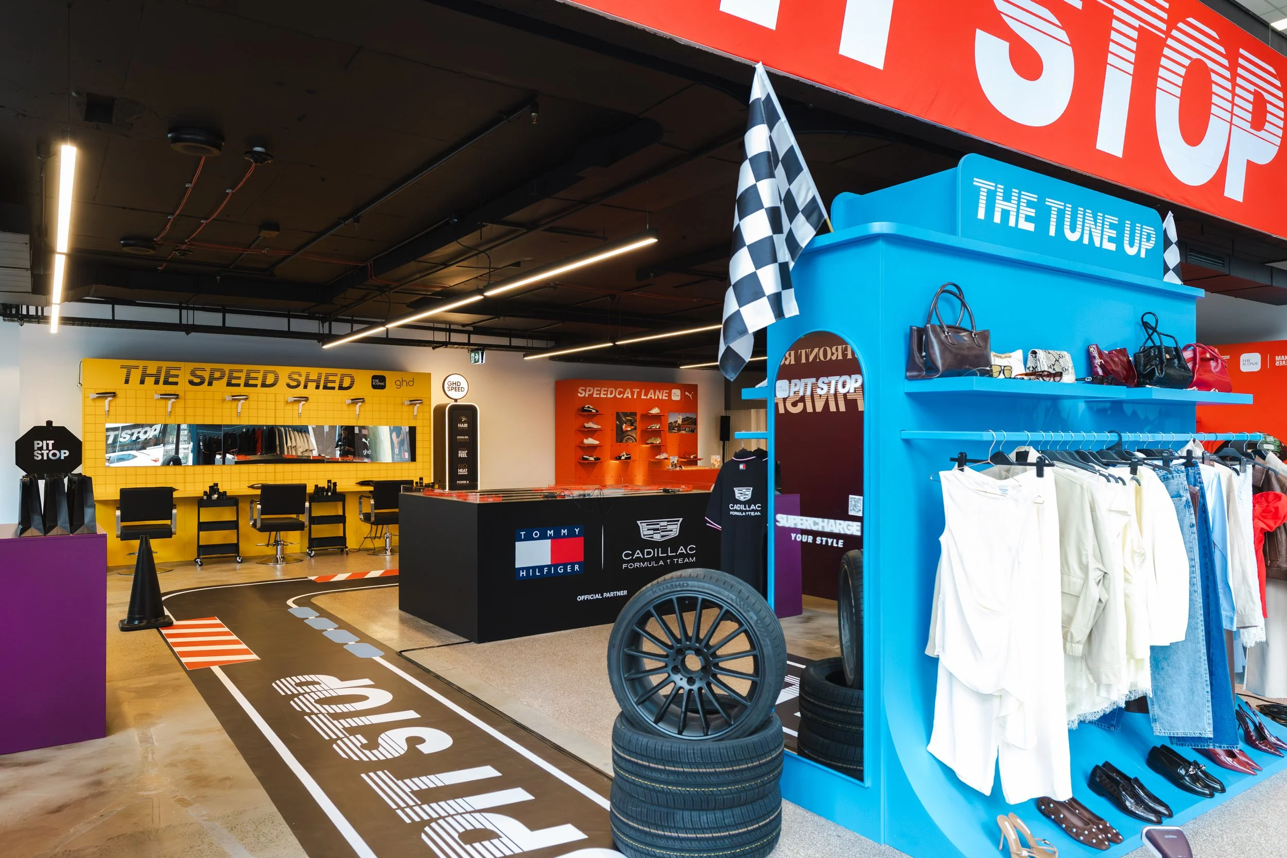 Interior of a shoe and accessory store featuring racing-themed displays, with tires on the floor and a large red 'Stop' sign overhead, along with various handbags, clothing, and branded signs.