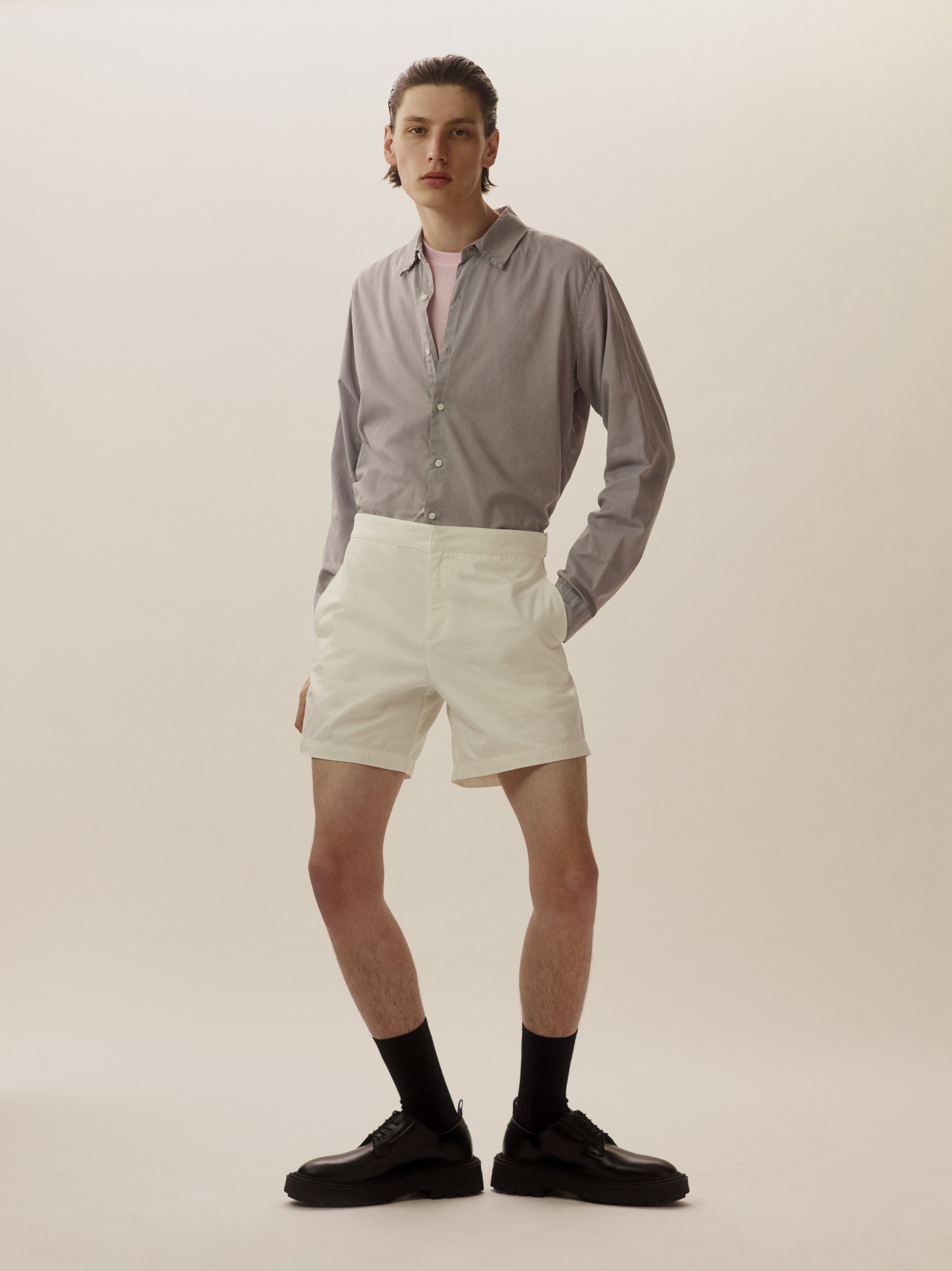 Young man standing with hands in pockets, wearing a gray button-up shirt, beige shorts, black socks, and black shoes, against a plain light background.
