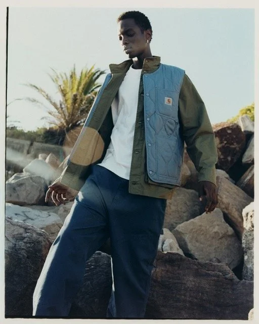 A young man in casual outdoor clothing, standing among rocks with a plant and clear sky in the background.