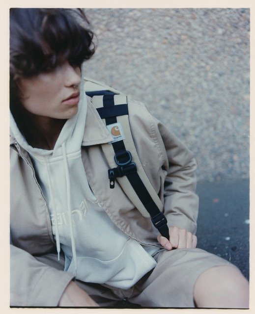 A young person with dark, curly hair sitting outdoors, wearing a beige jacket over a white hoodie, with a backpack strap over their shoulder.