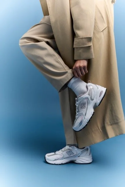 Person in beige coat and pants, wearing white sneakers, standing against a blue background.