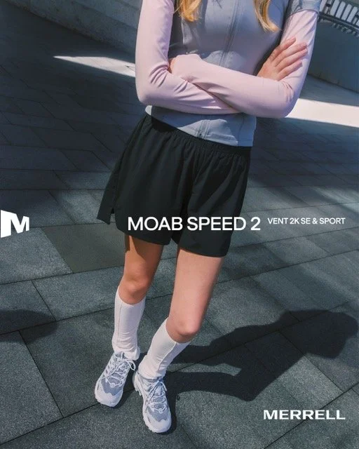 A woman stands outdoors with her arms crossed, wearing a light gray jacket, black athletic shorts, white socks, and gray running shoes.