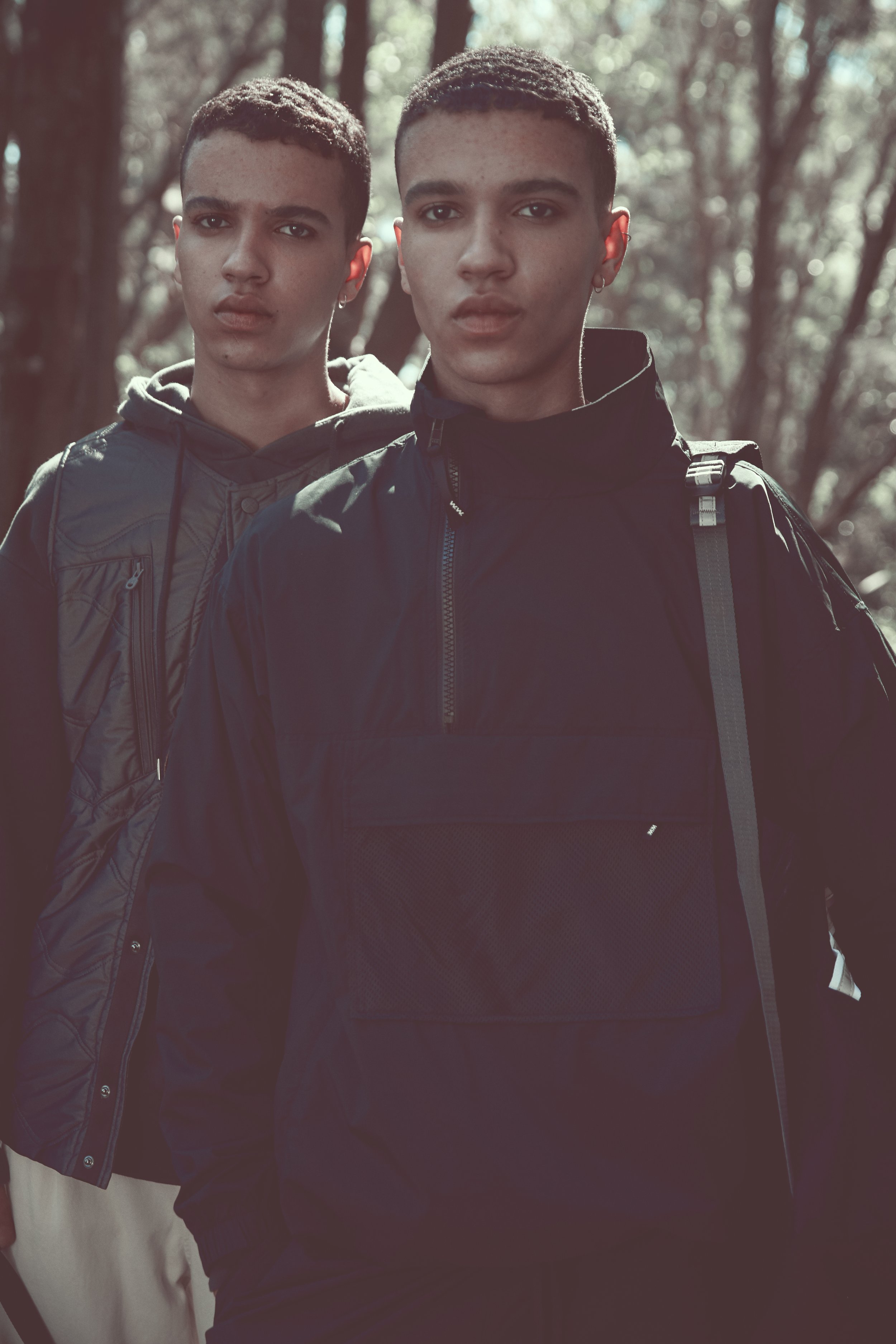 Two young men with short hair in a forested area, wearing dark jackets, standing close together and looking at the camera.