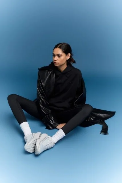 Young woman sitting on the floor against a blue background, dressed in a black hoodie, shiny black jacket, black leggings, white socks, and white sneakers.