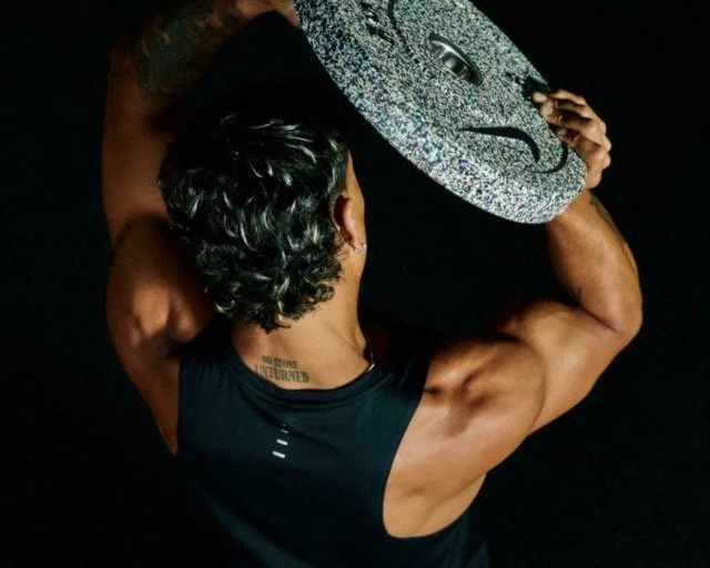 A muscular person lifting a large, glittery gray weight plate overhead, viewed from behind against a black background.
