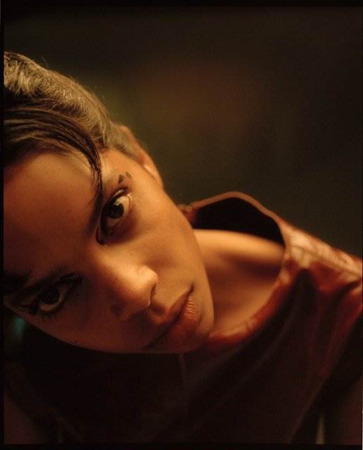 Close-up portrait of a young woman with dark hair and brown eyes, looking up at the camera, wearing a reddish-brown jacket, with a dark background.