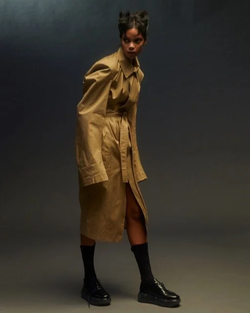 A woman wearing a tan trench coat, black socks, and platform boots standing against a dark background.