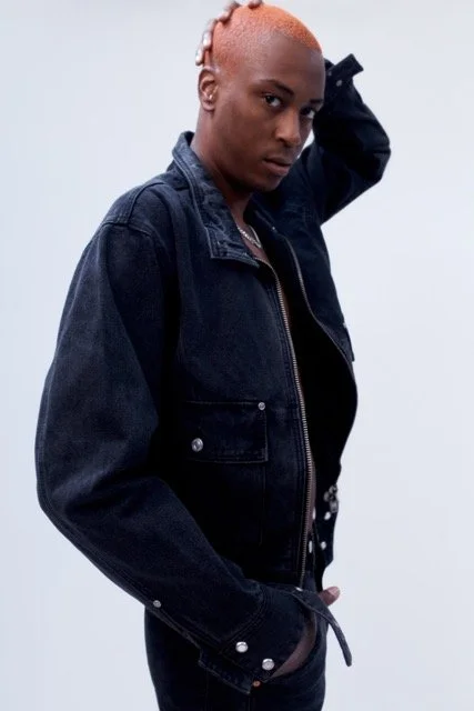Person with short, orange hair wearing a dark denim jacket and black shirt, posing with one hand on their head against a plain background.