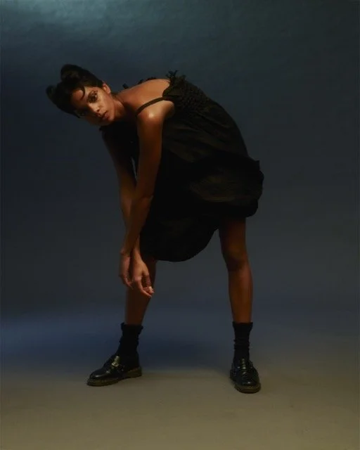 A woman in a black dress bending forward with one hand reaching toward the ground, standing against a dark background.
