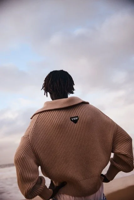 A person with short dreadlocks standing on a beach, facing the ocean and sky, wearing a tan oversized sweater.