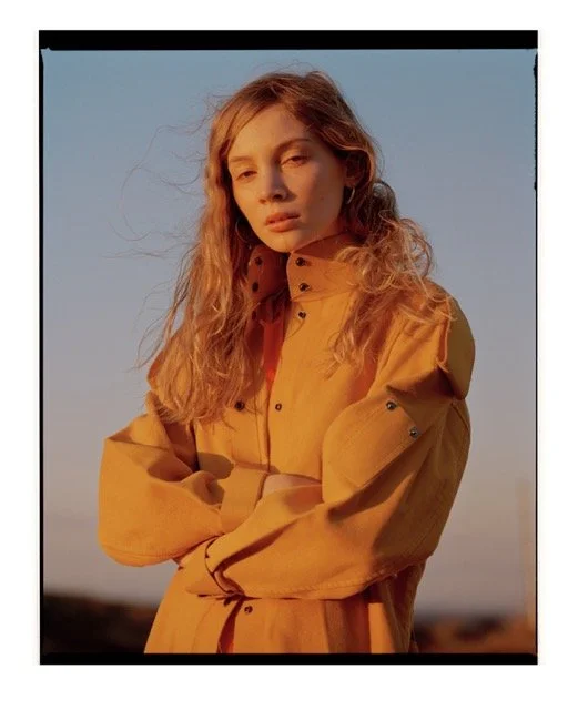 A young woman with long, wavy blonde hair wearing a loose, tan-colored jacket outdoors during golden hour.