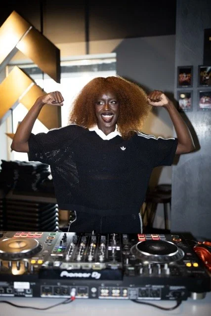 A woman with curly red hair flexing her biceps and smiling behind DJ equipment in a modern indoor setting.