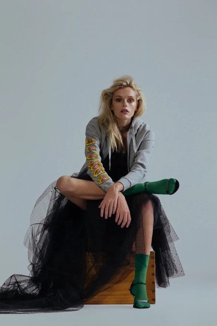 Young woman with blond hair sitting on a wooden box, wearing a gray hoodie, black tulle skirt, green socks, and high-heeled shoes, with a green microphone resting on her lap, against a plain gray background.