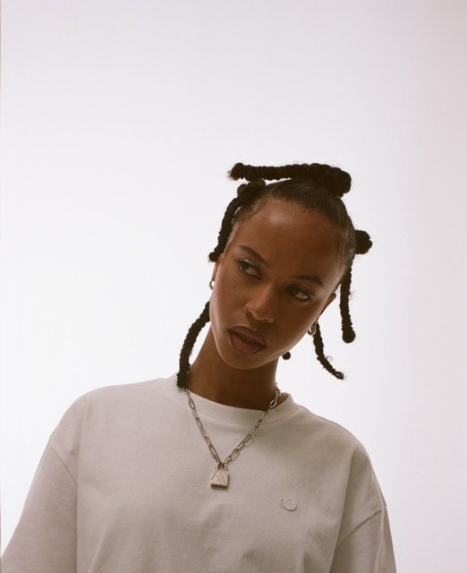 A woman with styled dreadlocks and dark lipstick wearing a white t-shirt with a small logo, a silver chain necklace, and earrings, looking slightly to the side against a plain background.