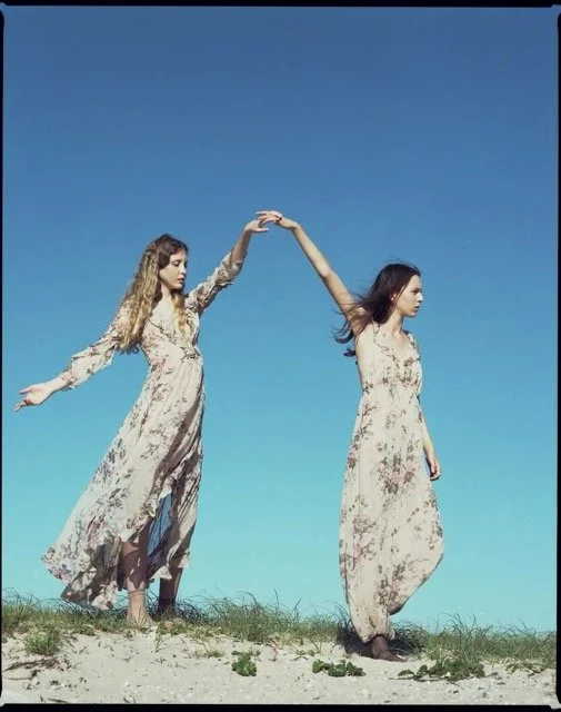Two women in long floral dresses standing on a grassy field under a clear blue sky, holding hands above their heads.