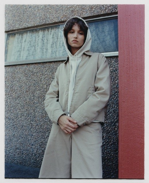 A young person with dark hair wearing a beige hoodie and jacket standing against a textured wall with a window and a red column.