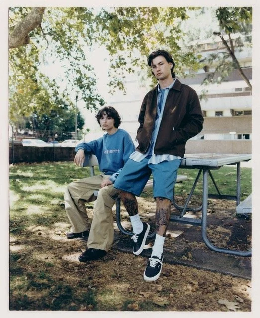 Two young men in casual clothing are outdoors in a park, sitting and standing near a picnic table, with trees and buildings in the background.