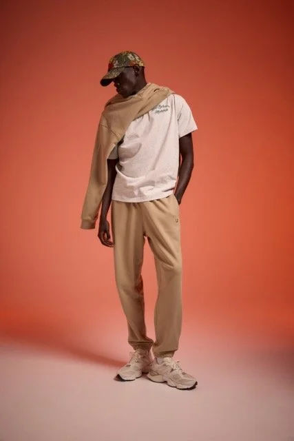 A man wearing a white T-shirt, beige sweatpants, sneakers, a camouflage baseball cap, and a tan jacket draped over his shoulders, standing against a pink background.