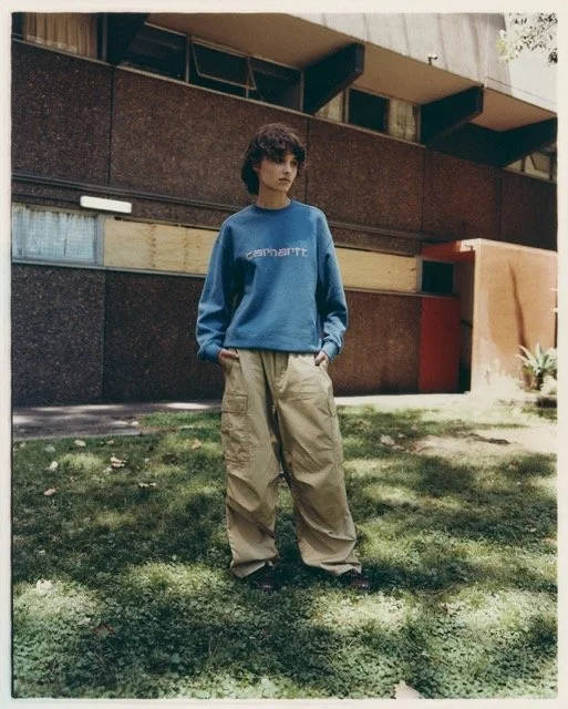 A young woman standing on a grassy area outside a building, wearing a blue sweatshirt and beige baggy pants, with her hands in her pockets.