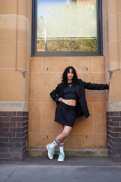 Young woman with curly hair wearing a crop top, black jacket, and skirt, standing against a brick and stone wall outside with one arm extended to the side and the other on her waist.