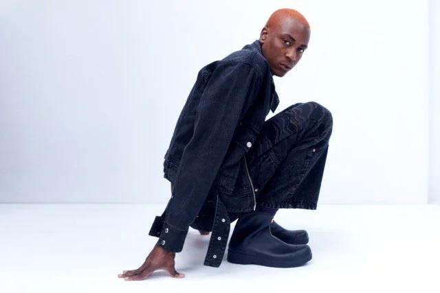 Person crouching on the floor in a black denim jacket, dark pants, and black shoes against a white background.