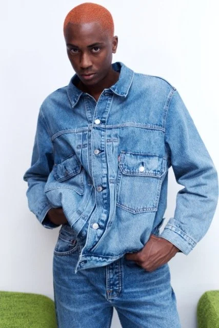 Person with short, orange hair wearing a blue denim jacket and jeans, standing against a white background.