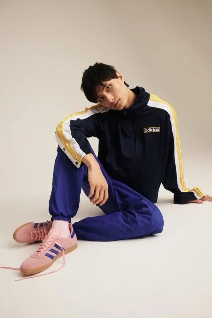 A young man dressed in a navy and gold Adidas sports tracksuit, sitting on the floor with one knee raised and one arm resting on it, against a plain beige background.