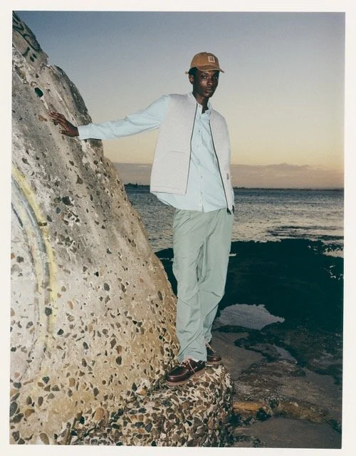 A man standing on a rocky shoreline at sunset, wearing a baseball cap, a light jacket, and light-colored pants, with his hand resting on a large concrete structure.