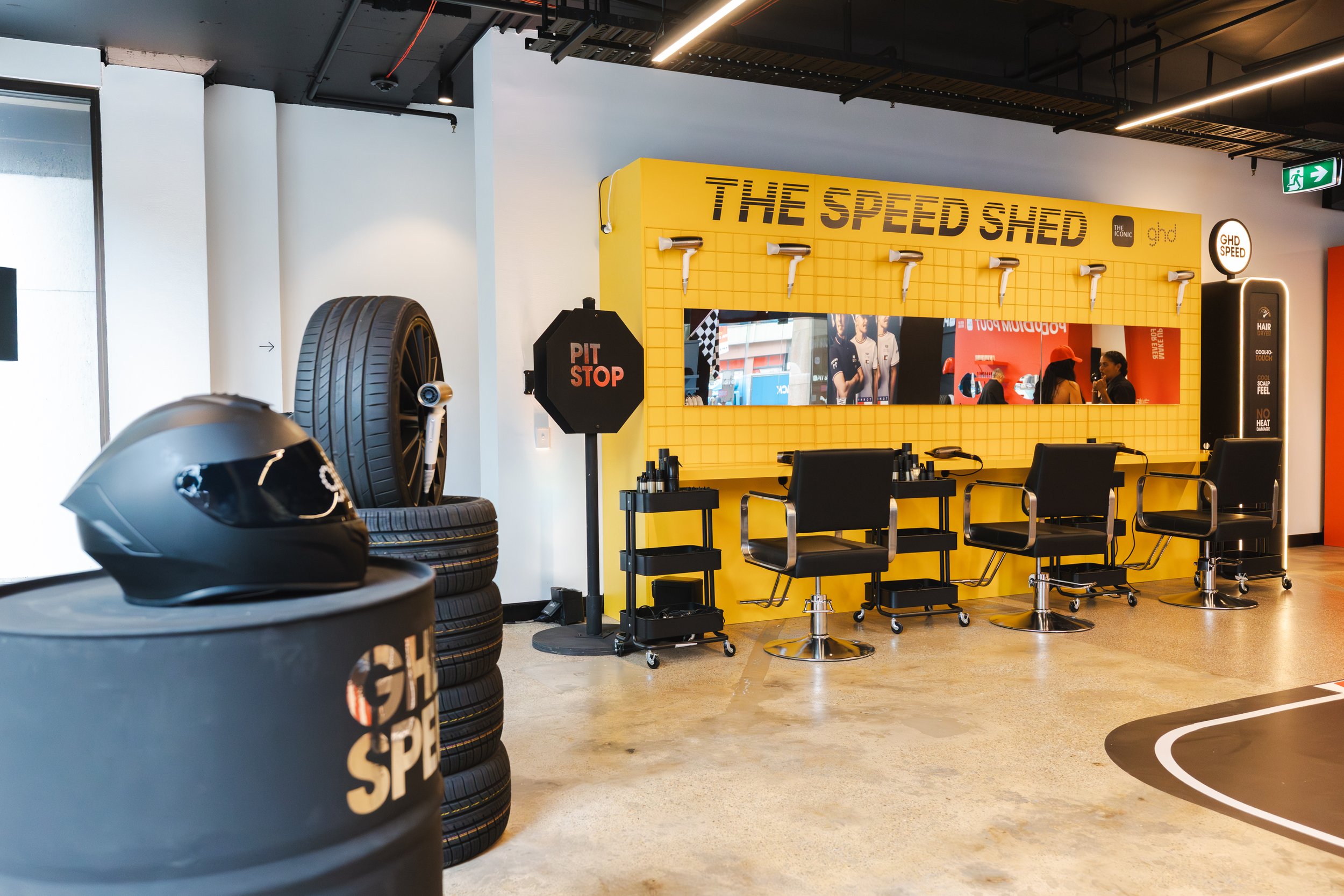 The image shows the interior of a hair salon or barbershop themed as a race car pit stop. There are tires stacked on the left, a helmet on top of the tires, and a yellow wall with the words 'THE SPEED SHED' and hair dryers mounted on it. Chairs and hairdressing stations are lined up in front of a mirror, with a few people visible in the reflection. An exit sign is visible on the upper right.