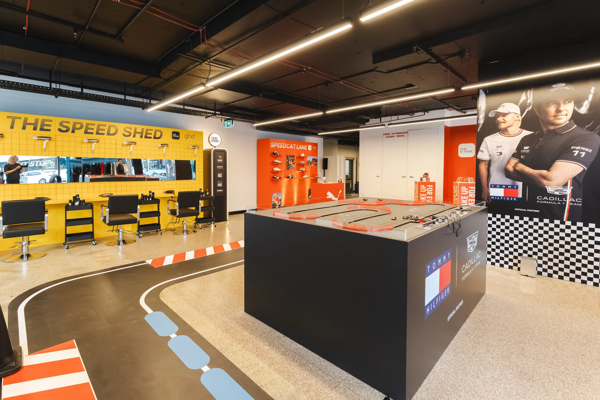 Indoor slot car racing track with a black surface, blue and white track markings, and a surrounding yellow wall labeled 'The Speed Shed'. There are chairs and mirrors on the yellow wall, and a large poster of two men in racing attire on the right side. Overhead lights illuminate the space.