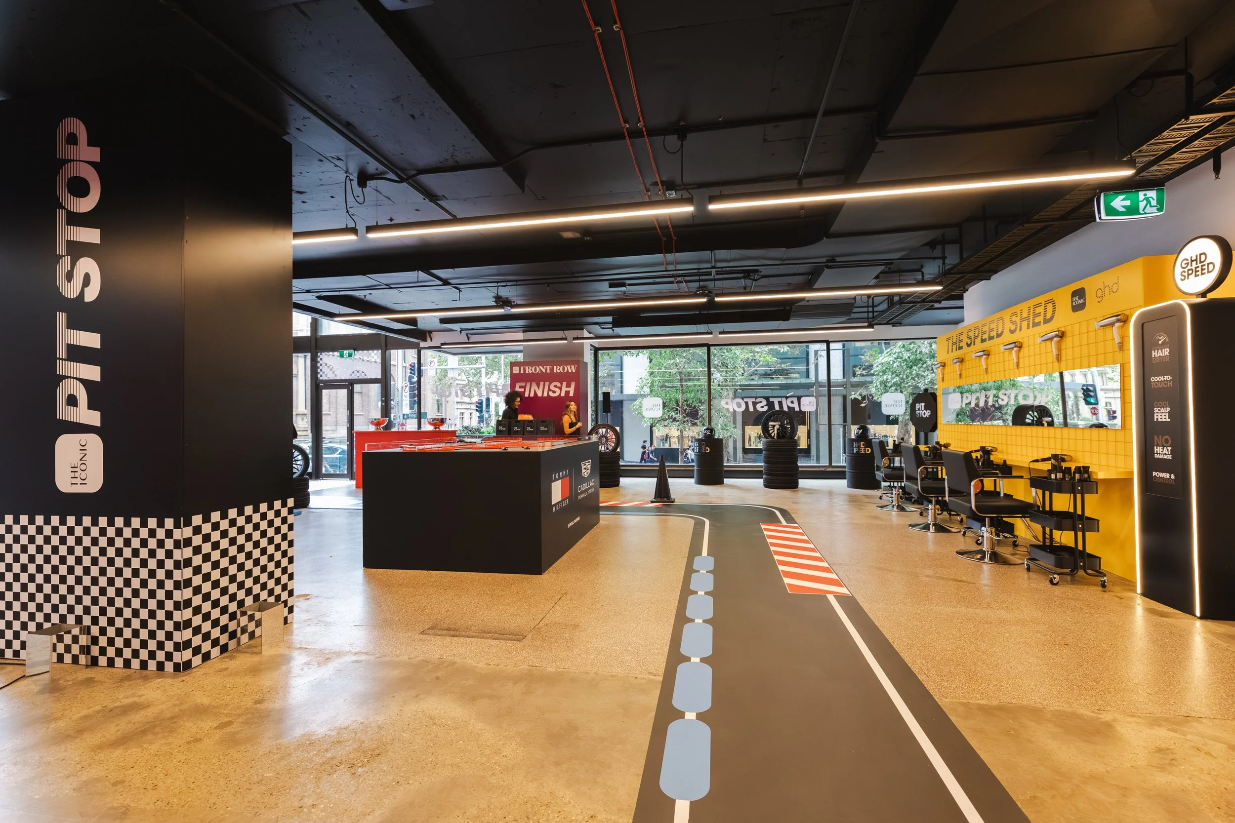 Inside a modern car repair or detailing shop with signage for 'The Speed Shed' and 'Pit Stop,' featuring a designated area for vehicle maintenance, a 'Finish' sign at the front, black tires stacked, chairs, and a woman working at a reception desk near the large front windows with trees outside and a partly cloudy sky.