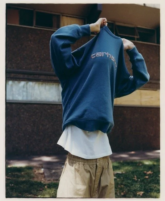 Person standing outdoors, pulling a blue sweatshirt over their face, wearing beige shorts and a white shirt underneath.