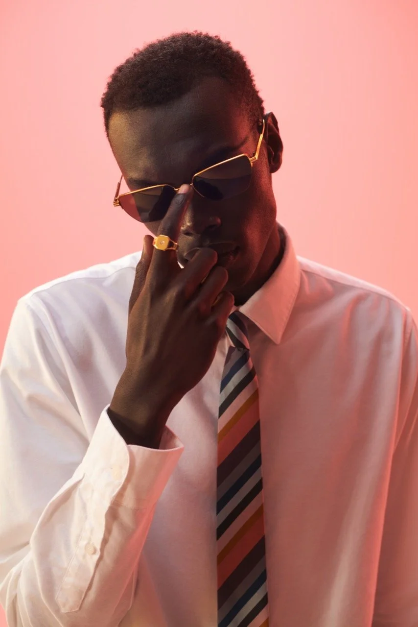 A young man with dark skin wearing sunglasses, a white dress shirt, and a striped tie, has one hand raised to his face touching his sunglasses, against a pink background.