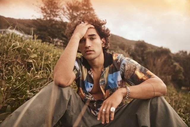 A young man sitting outdoors on grass with a sunset sky in the background, resting his head on his hand and looking at the camera.