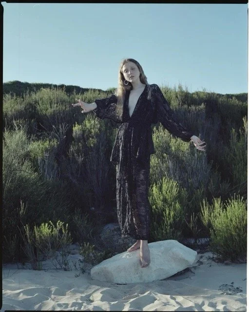A woman in a black dress standing barefoot on a rock in a sandy area with greenery behind her.