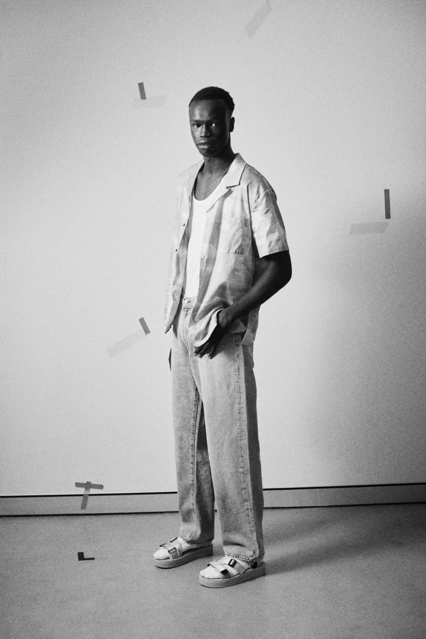 Black and white photo of a young man standing against a plain wall, wearing casual clothing with hands in pockets, looking directly at the camera.