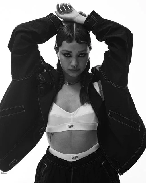 Black and white photo of a young woman with dark hair styled in braids, wearing a sports bra and an oversized jacket, looking directly at the camera with her arms raised behind her head.
