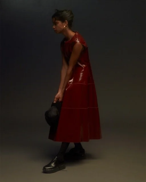 A woman with short hair in a red vinyl dress, black boots, and earrings, tilting forward holding a black hat against a dark background.