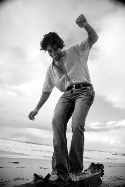 Person standing on a surfboard on the beach, smiling and raising one fist in the air, with the ocean and cloudy sky in the background.