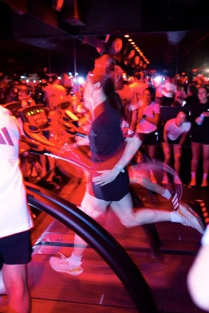 A woman jumping with her knees bent and arms outstretched in a lively, crowded nightclub with colorful lights.