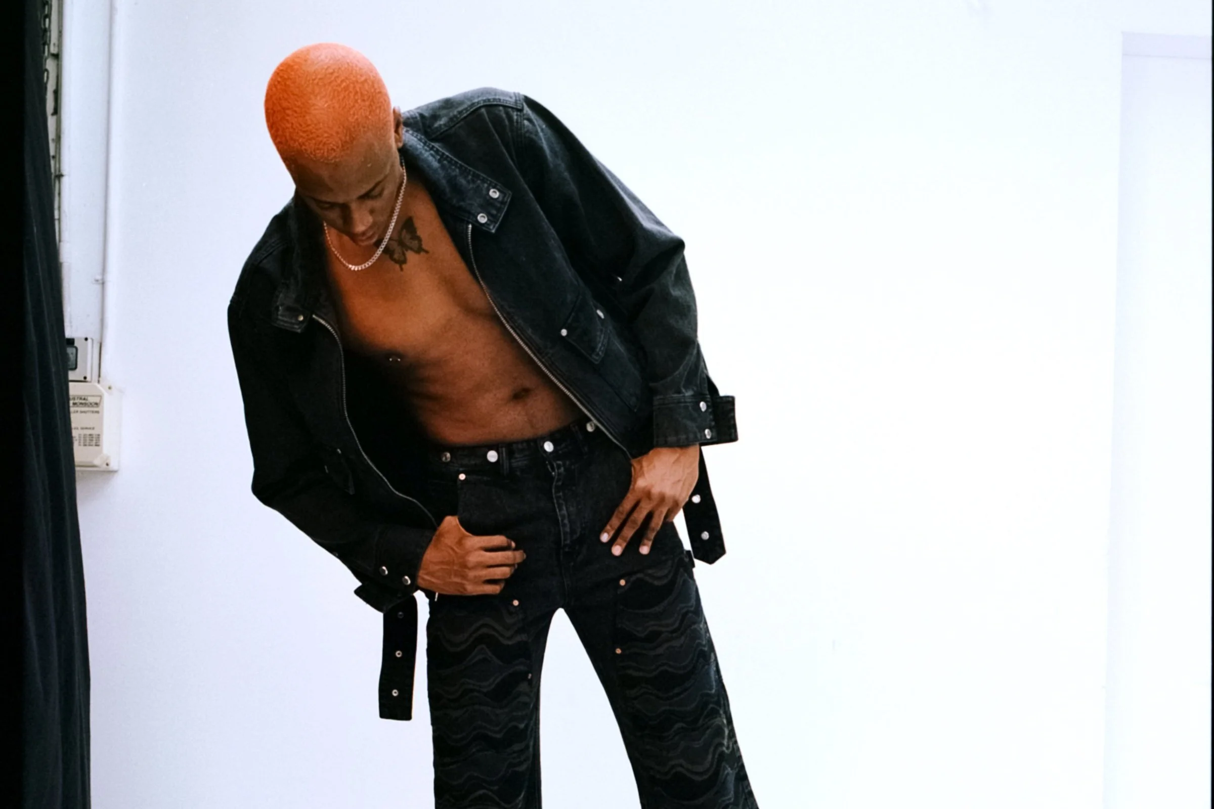 A person with short orange hair wearing a black denim jacket and black pants, standing against a plain white background, looking down and with one hand on his hip.