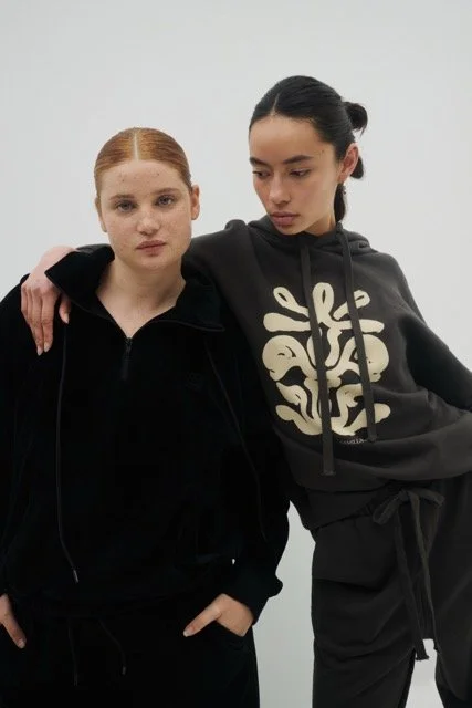 Two young women standing together against a plain white background, one with red hair and freckles wearing a black zip-up hoodie, the other with dark hair in a bun wearing a black hoodie with a beige graphic design.